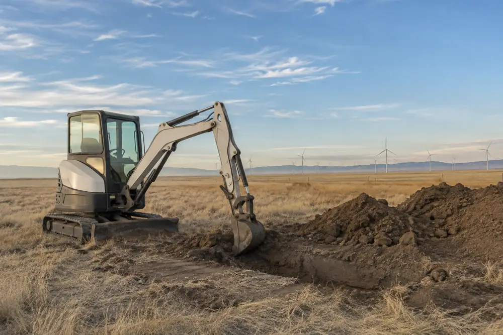 Mini excavator at a fresh pit on flat high-plains acreage with wind turbines in the distance